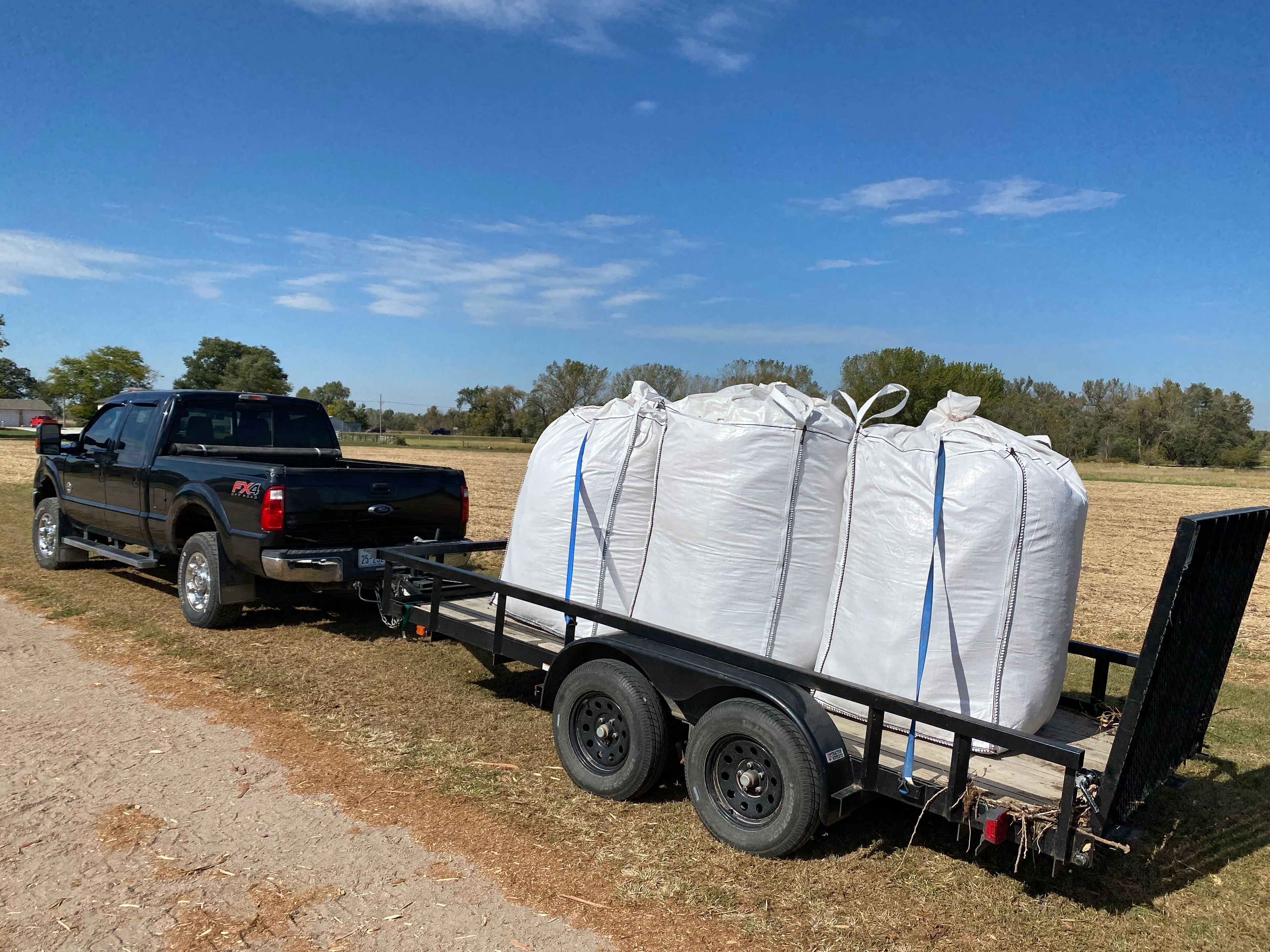 Black pickup truck with a trailer carrying large white bags of Cedar barons show fiber on a rural road.
