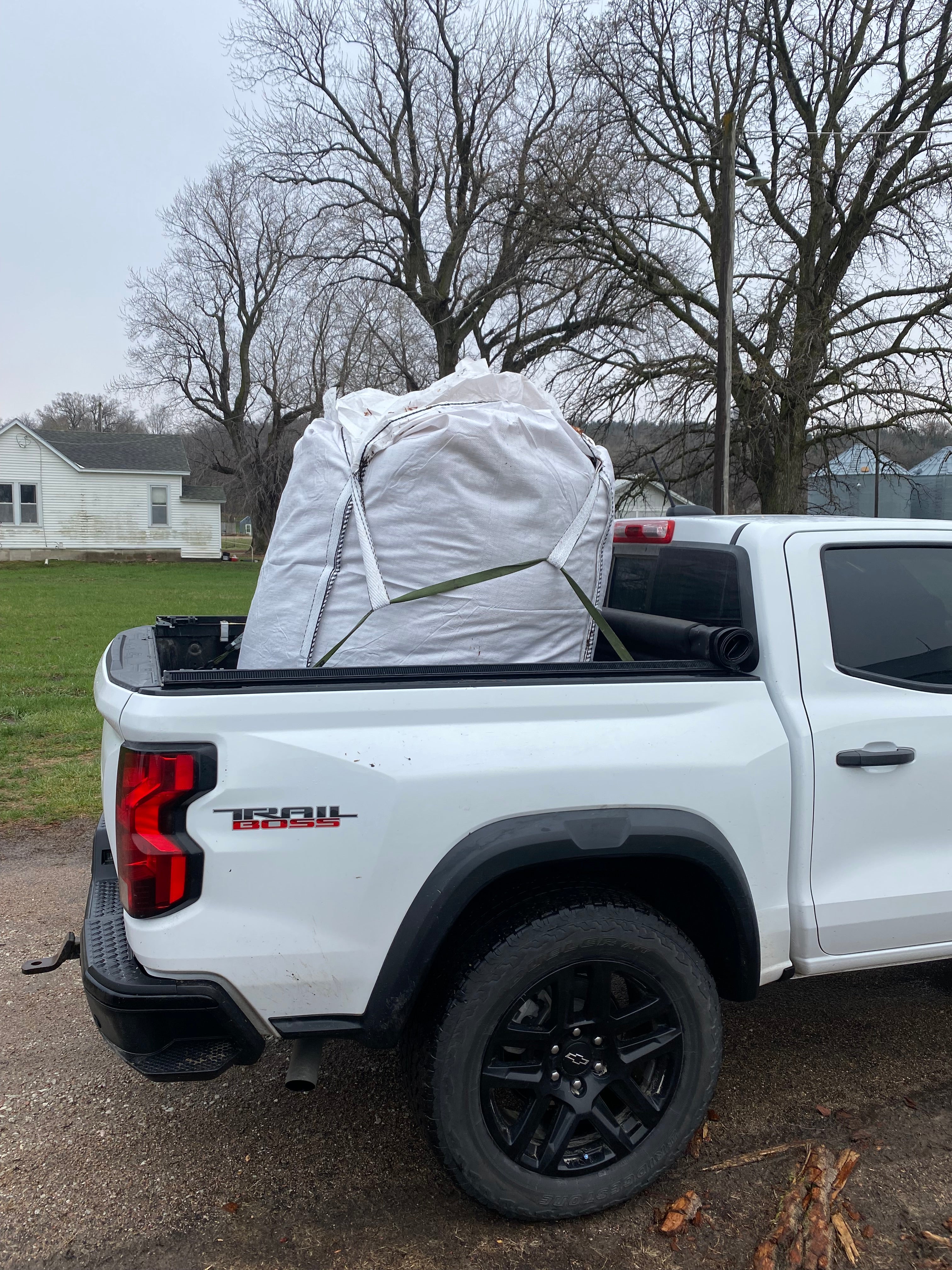 White truck with a large white bag in the bed, parked on a dirt road with leafless trees in the background. Cedar Barons Show Fiber