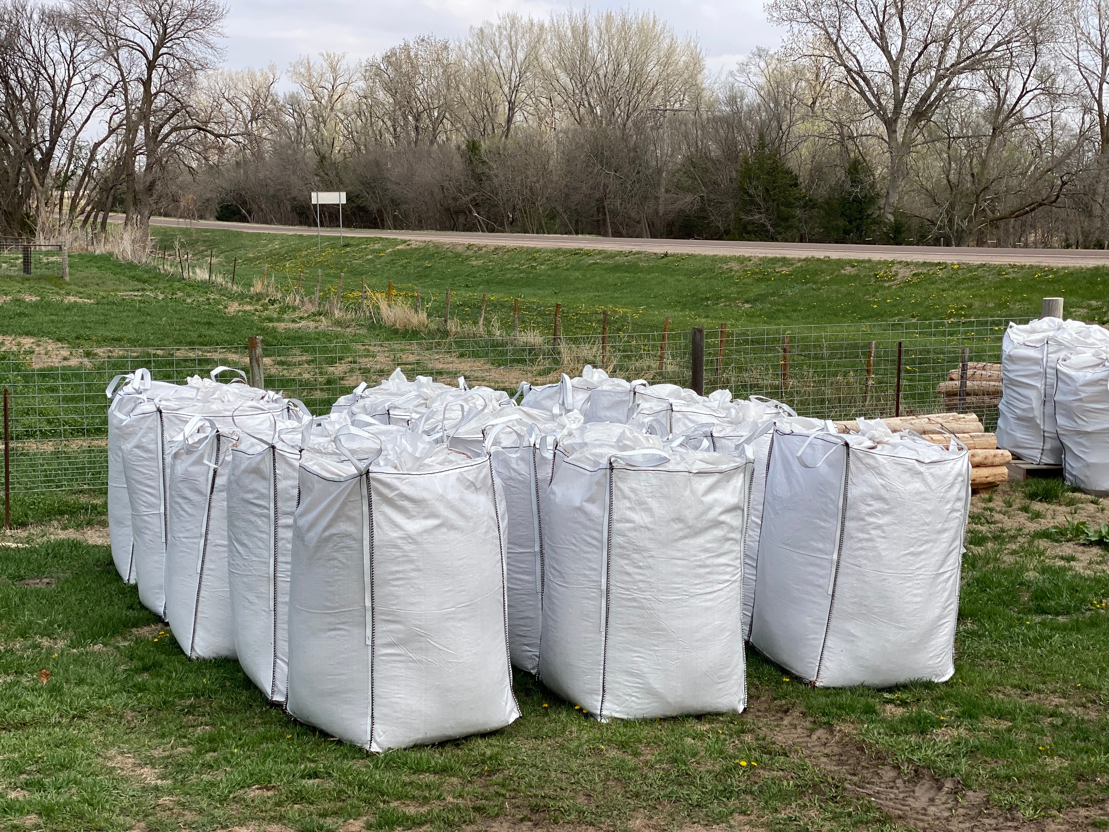 Large white bags on a grassy field with trees and a road in the background Cedar Barons Show Fiber 
