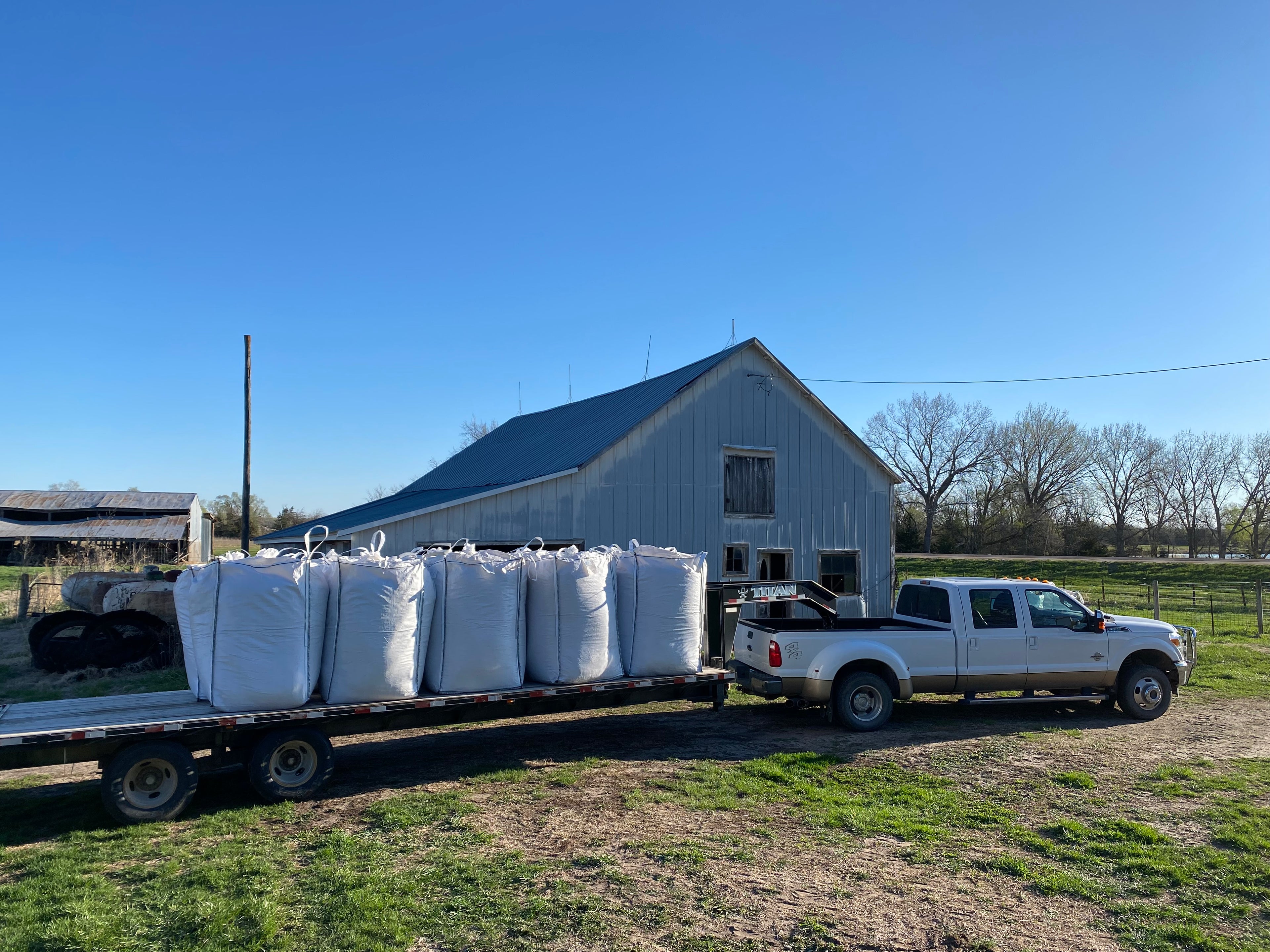 White truck with a trailer loaded with large white bags of Cedar Barons Show Fiber in front of a barn on a clear day.